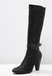 Botte en cuir noir hauteur genou avec un bout pointu, une surface texturée et un détail de sangle à la cheville avec une boucle. Design à talon aiguille.