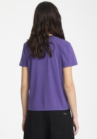 Maglietta di cotone viola a maniche corte, con scollo rotondo e orlo dritto, indossata con jeans neri a vita alta con tasche posteriori.