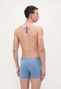 Kék férfi boxeralsó, puha anyagból, szürke derékkal, amelyen a "Copenhagen Studios" logó található. A modellen tetoválás látható a hátán.