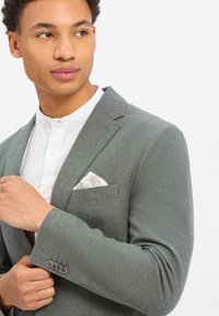Grijze blazer van gestructureerde stof, met een diepe revers, vier knoopdetails op de manchetten en een witte met patroon pocket square.