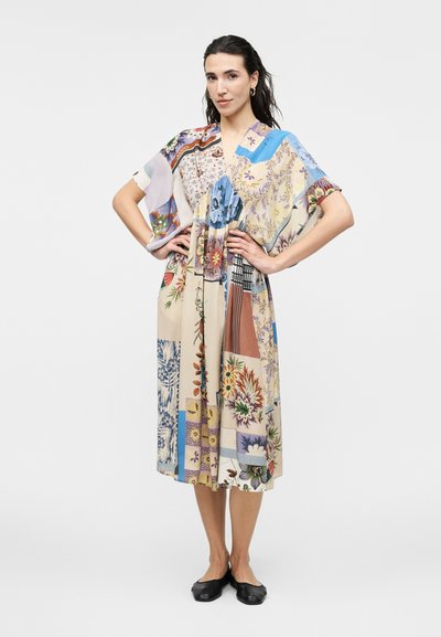 Femme debout, mains sur les hanches, portant une robe midi fleurie patchwork colorée et des chaussures plates noires sur un fond uni.