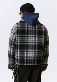 Persoon in zwarte beanie, bril, zwart-grauwe geruite jas met marineblauwe capuchon en zwarte broek met kettingaccessoires, gezien vanuit de achterkant.