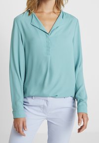 Blouse turquoise à manches longues en tissu lisse avec col rabattu et patte de boutonnage à l'avant, associée à un pantalon bleu clair.