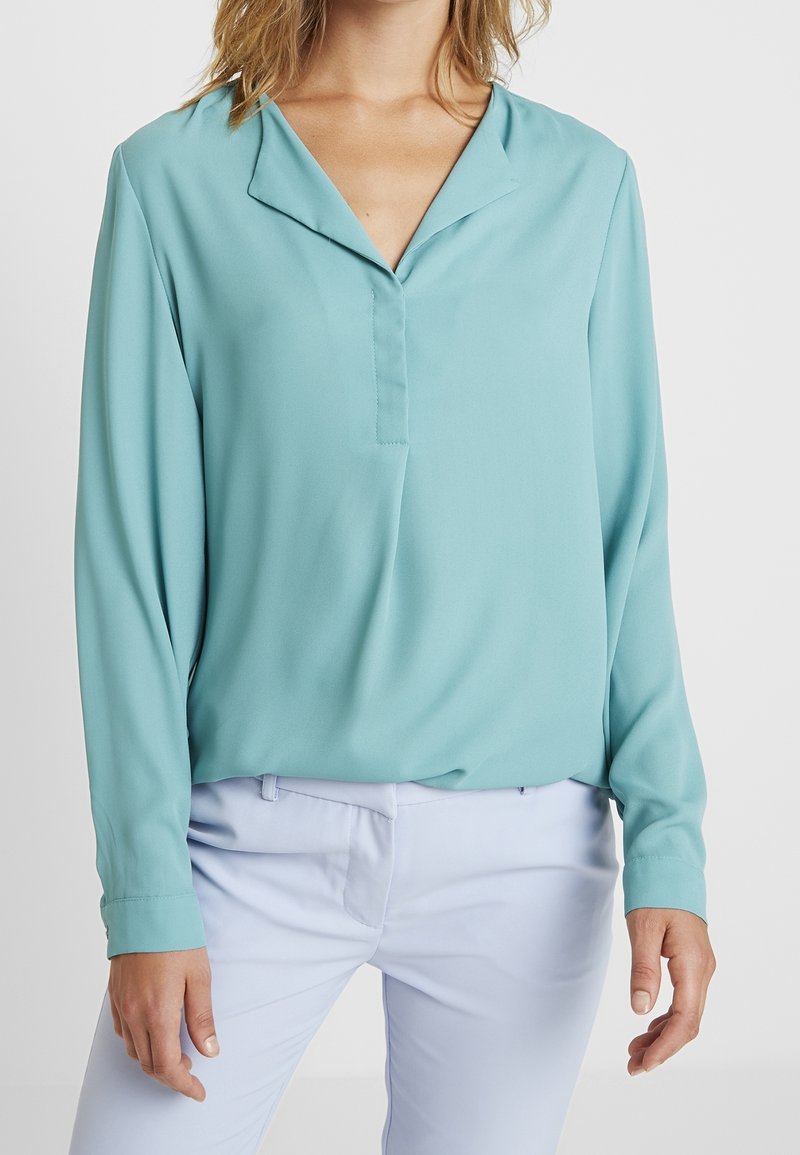 Blouse turquoise à manches longues en tissu lisse avec col rabattu et patte de boutonnage à l'avant, associée à un pantalon bleu clair.