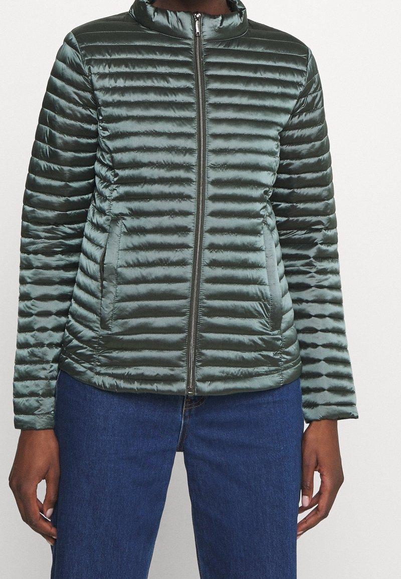Personne portant une veste matelassée verte avec une fermeture éclair frontale et des poches, associée à un jean bleu en denim, visible des épaules aux hanches.
