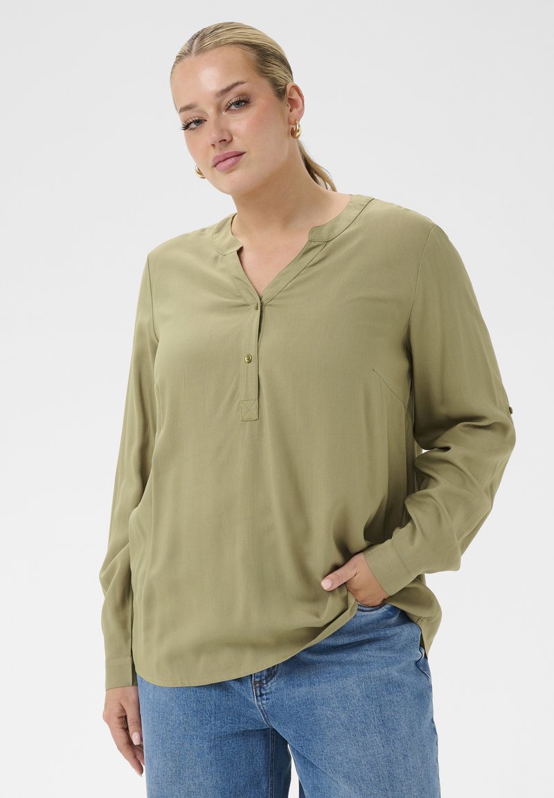 Femme portant un chemisier manches longues vert olive avec un col boutonné et un jean bleu, debout avec une main dans la poche devant un fond uni.