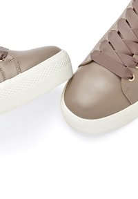 Beige synthetic leather sneakers with a smooth finish, featuring light brown laces, gold eyelets, and white rubber soles with textured patterns.