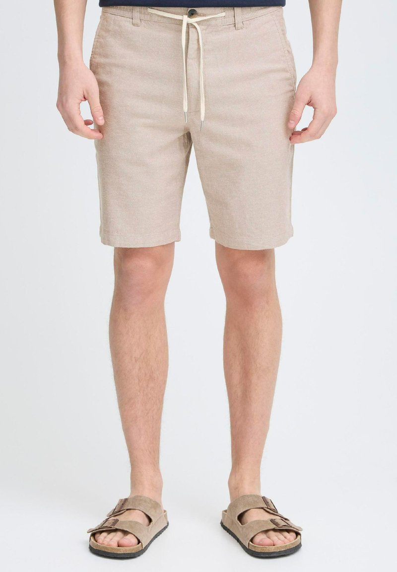 Shorts beige taillés en tissu texturé, avec cordon de serrage et fermeture à bouton, ourlets roulés, associés à des sandales marron clair.