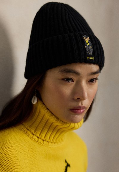 Melna ribota adīta cepure ar logo uzlīmi, kurā attēlots lācis uzvalkā. Valkāta kopā ar dzeltenu augstu apkakli džemperi, rādot teksturētu adījumu.