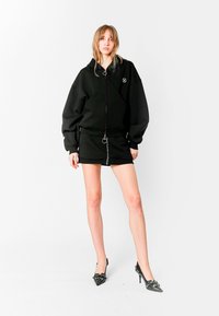 Sudadera negra de gran tamaño con cierre de cremallera y detalle de logo, combinada con una falda mini negra con cremallera frontal. Tacones negros de punta con adornos.