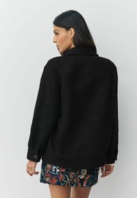 Femeie cu păr lung și închis la culoare, purtând o jachetă neagră texturată și o fustă scurtă cu imprimeu floral, stând în picioare lângă un perete simplu, deschis la culoare, cu profilul orientat într-o parte.