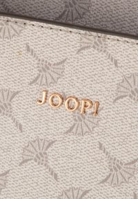 Wit textuurmateriaal met een subtiel patroon. Goudfolie "JOOP!"-logo centraal en opvallend weergegeven.