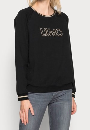 Woman wearing a black Liu Jo sweatshirt with gold trim on neckline and cuffs, paired with gray jeans, standing against a plain background.