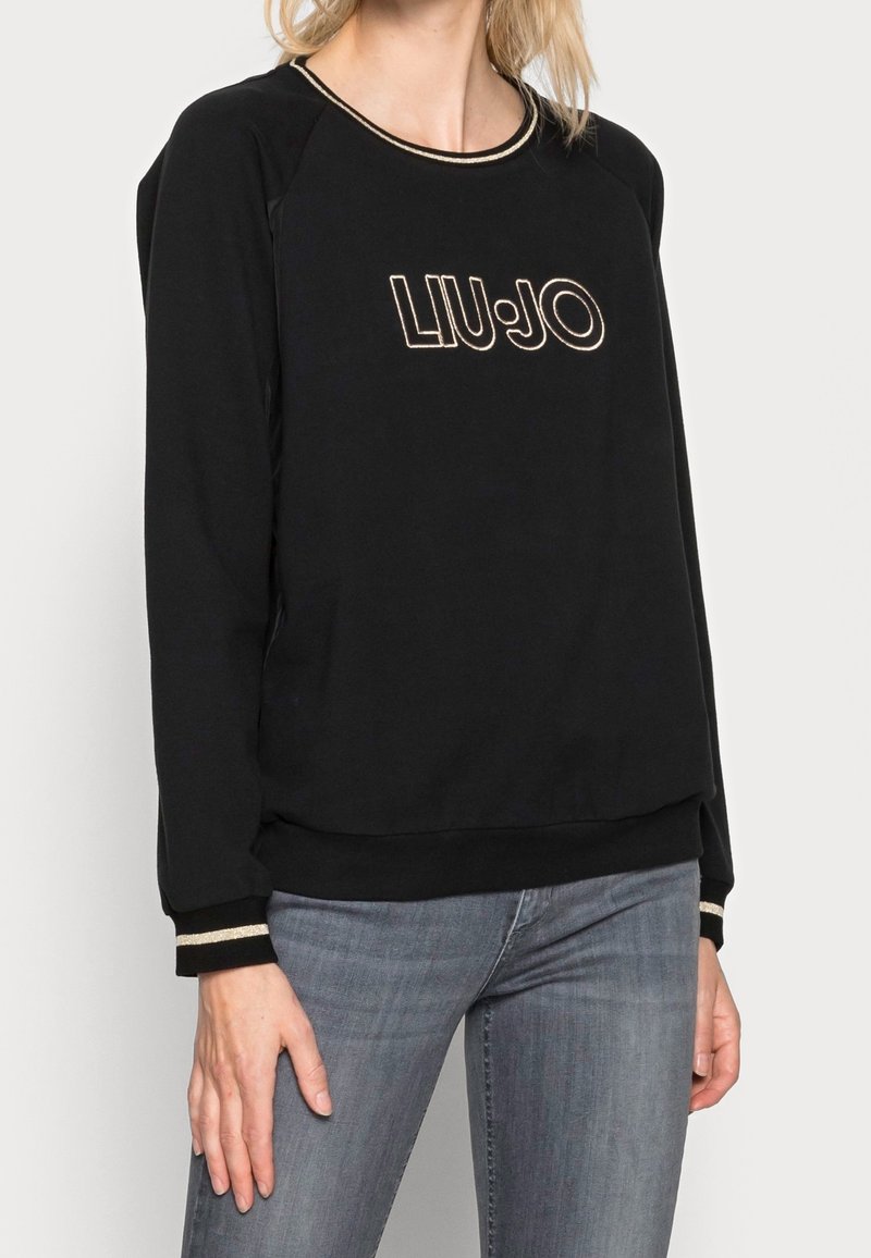 Femme portant un sweatshirt Liu Jo noir avec des bordures dorées au niveau de l'encolure et des poignets, associée à un jean gris, debout devant un fond uni.
