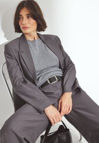 Pilkos pritaikytos kelnės su plačiais sijonais, tekstūruotas pilkas viršus ir juodas diržas. Juoda rankinė stovi ant žemės, pasvirus prie sidabrinės kėdės.