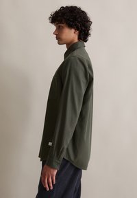 Giovane uomo con capelli ricci che indossa una camicia a maniche lunghe verde oliva e pantaloni scuri, in piedi di profilo su uno sfondo semplice.