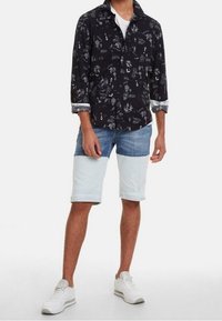 Camisa negra abotonada con estampado gráfico blanco, combinada con pantalones cortos de mezclilla azul claro. El modelo lleva zapatillas blancas. Los pantalones cortos tienen un patrón combinado por bloques de color.