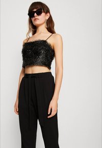 Kvinna i svart texturerad ärmlös crop top, högmidjade svarta byxor med bälte, stora hoops örhängen och svarta överdimensionerade solglasögon.