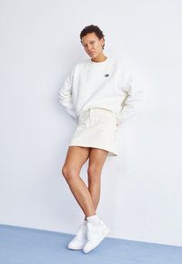 Valkoinen pusero logolla, beige hame ja valkoiset lenkkarit. Asukokonaisuus on rento ja minimalistinen, vaalealla taustalla.