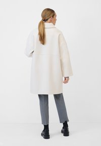 Weißer übergroßer Mantel mit Kragen, strukturiertem Stoff und sichtbaren Nähdetails. Kombiniert mit grauen maßgeschneiderten Hosen und schwarzen Stiefeln.