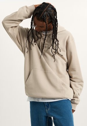 Sweatshirt à capuche beige avec une poche avant et un patch logo. Porté sur une chemise grise, associé à un jean bleu. Tissu texturé et coupe décontractée.