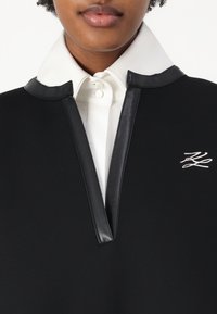 Juodas drabužis su baltu vidiniu apykakle, pasižymintis užtrauktuko kaklu, kontrastingais odiniais akcentais ir sidabriniu logotipo detaliu ant krūtinės.