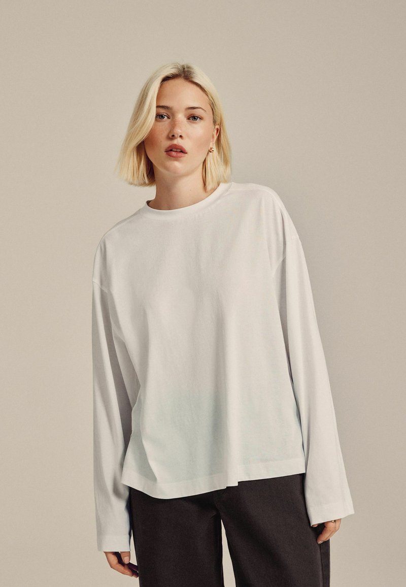 Witte longsleeve t-shirt met een relaxed fit, ronde halslijn en subtiele gradiënt aan de onderrand. Gecombineerd met donkere broeken.
