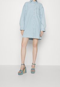 Abito camicia in denim azzurro chiaro con maniche lunghe e chiusura a bottoni, abbinato a sandali con plateau color teal e cinturini alla caviglia.