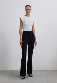 Weißes strukturiertes Oberteil mit kurzen Ärmeln, kombiniert mit hoch taillierten schwarzen Schlagjeans. Das Model trägt schwarze Sandalen vor einem hellgrauen Hintergrund.