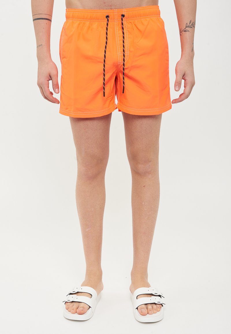 Pantaloni da bagno arancioni neon con vita elastica, cordoncino nero e cuciture bianche; indossati con sandali bianchi.