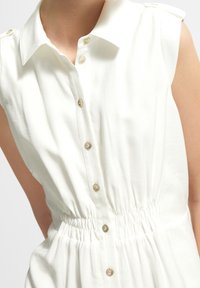 Robe blanche sans manches avec un devant à boutons, un col, une taille élastique et une texture de tissu lisse.