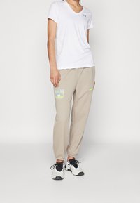 Weißes kurzärmliges V-Ausschnitt-T-Shirt, kombiniert mit beigen Jogginghosen mit einem bunten Grafikdesign und einem neongrünen Akzent auf der Tasche. Schwarze Sportschuhe.