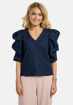 Femme souriante aux cheveux blonds portant un haut bleu foncé à manches bouffantes et un pantalon rose clair sur un fond uni.