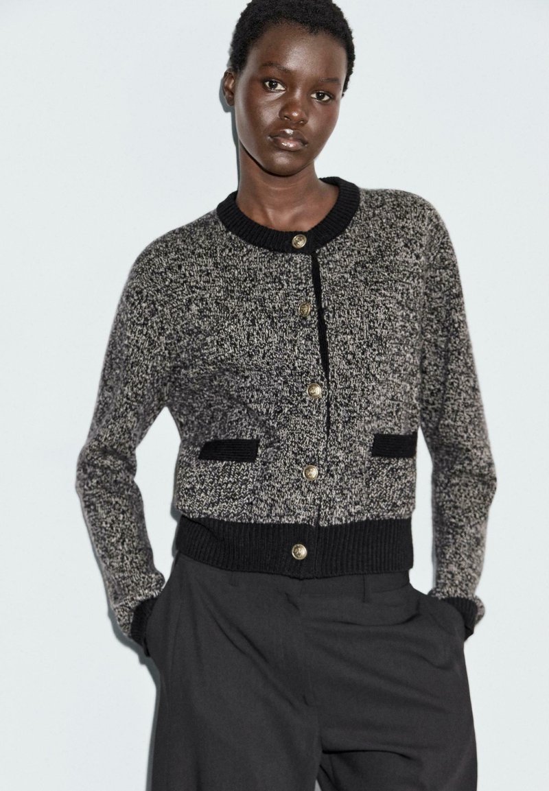 Cardigan textures in bianco e nero con colletto tondo, bottoni dorati e dettagli neri sulle tasche. Indossato sopra pantaloni scuri.
