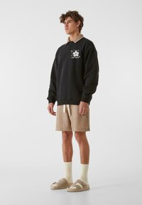 Melns krekls ar baltu ziedu rakstu un bēša krāsas apkakli, brūni īsie džinsi ar tekstūru, krēms krāsas zeķes un brūnas slip-on kurpes ar siksnām.