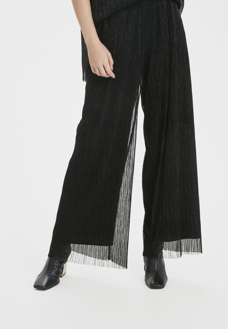 Pantalon large noir transparent porté avec des bottines noires et un haut assorti, montré de mi-cuisse aux pieds sur un fond blanc.