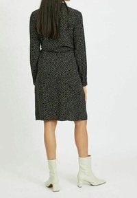 Žena v černých šatech po kolena s bílými skvrnami a bílými kozačkami na středním podpatku, stojící proti jednoduchému bílému pozadí.