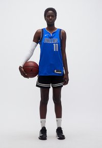 Γυναίκα παίκτρια μπάσκετ με μπλε φανέλα των Dallas κρατάει μπάλα μπάσκετ, στέκεται μπροστά σε λευκό φόντο.