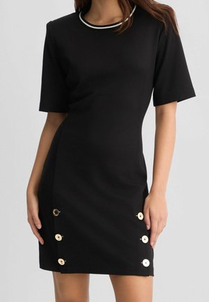 Femme portant une courte robe noire à manches courtes, un col bordé de blanc et six boutons décoratifs près de l’ourlet de chaque côté.
