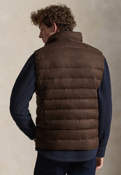 Mężczyzna o kręconych włosach, ubrany w brązową pikowaną kamizelkę na granatowej polarowej kurtce, zwrócony tyłem na jednolitym tle.