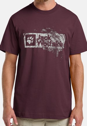 Mand iført en vinrød T-shirt med et slidt hvidt Jack Wolfskin-logo og poteaftryk på brystet.