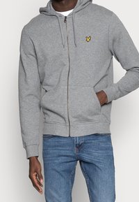 Grauer Reißverschluss-Hoodie aus weichem Material, mit Kapuze, Kängurutaschen und einem kleinen gelben Logo auf der linken Brust. Getragen mit blauen Jeans.