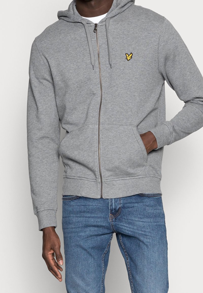Grauer Reißverschluss-Hoodie aus weichem Material, mit Kapuze, Kängurutaschen und einem kleinen gelben Logo auf der linken Brust. Getragen mit blauen Jeans.