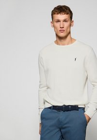 Suéter largo de manga larga color crema con cuello redondo, combinado con pantalones azules y un cinturón trenzado oscuro. Pequeño detalle de logo en el suéter.
