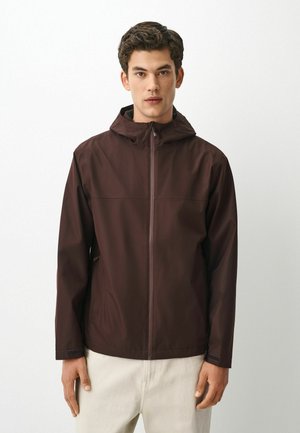 Young man wearing a dark brown hooded jacket with zipper, standing against a plain light background, facing forward.