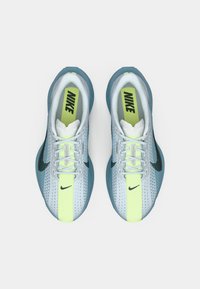 Zila un baltā Nike skriešanas apavu pāris ar neona dzeltenām valkājošajām lentēm un melniem Swoosh logotipiem, skatīts no augšas uz balta fona.