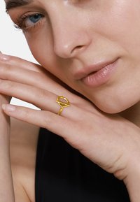 Anello d'oro con un design astratto caratterizzato da forme circolari e lineari. Indossato su una mano con smalto neutro su uno sfondo semplice.