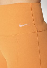 Nike legíny s vysokým pasem ve světlé oranžové látce. Mezi vlastnosti patří hladká textura, ploché švy a malé bílé logo na pase.