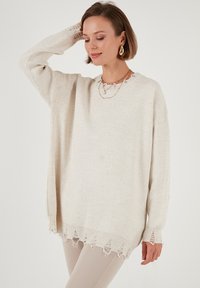 Maglione over-size color crema con colletto a coste e orlo sfrangiato. Tessuto morbido con dettagli intricati sui polsini.