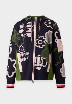 Femme aux longs cheveux noirs porte un pantalon noir à jambes larges, un haut noir et un sweat à capuche zippé noir avec de grands motifs floraux verts et blancs.